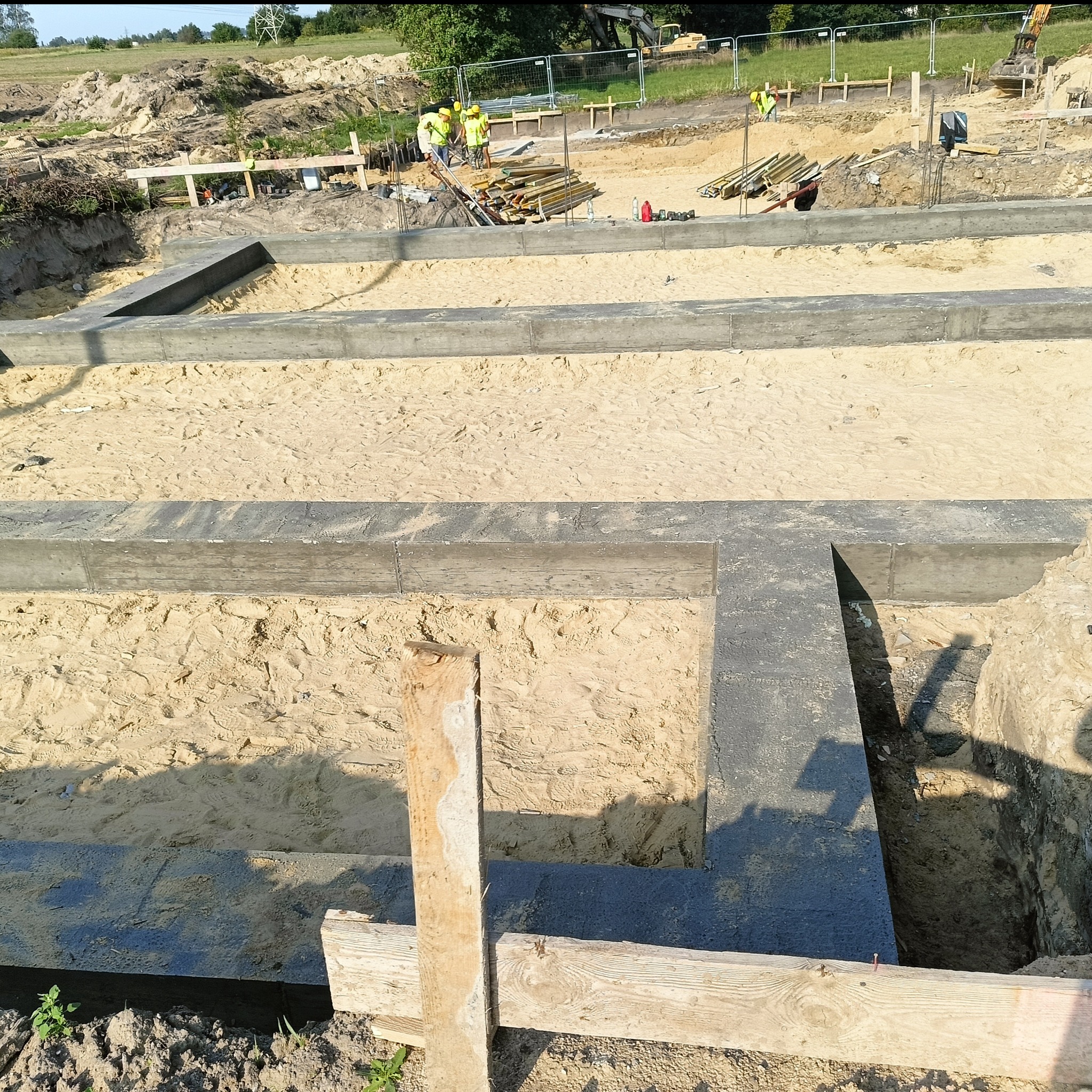 Zalane fundamenty domu jednorodzinnego z widocznym zbrojeniem i drewnianym szalunkiem. W tle ekipa budowlana w żółtych kamizelkach i ciężki sprzęt.