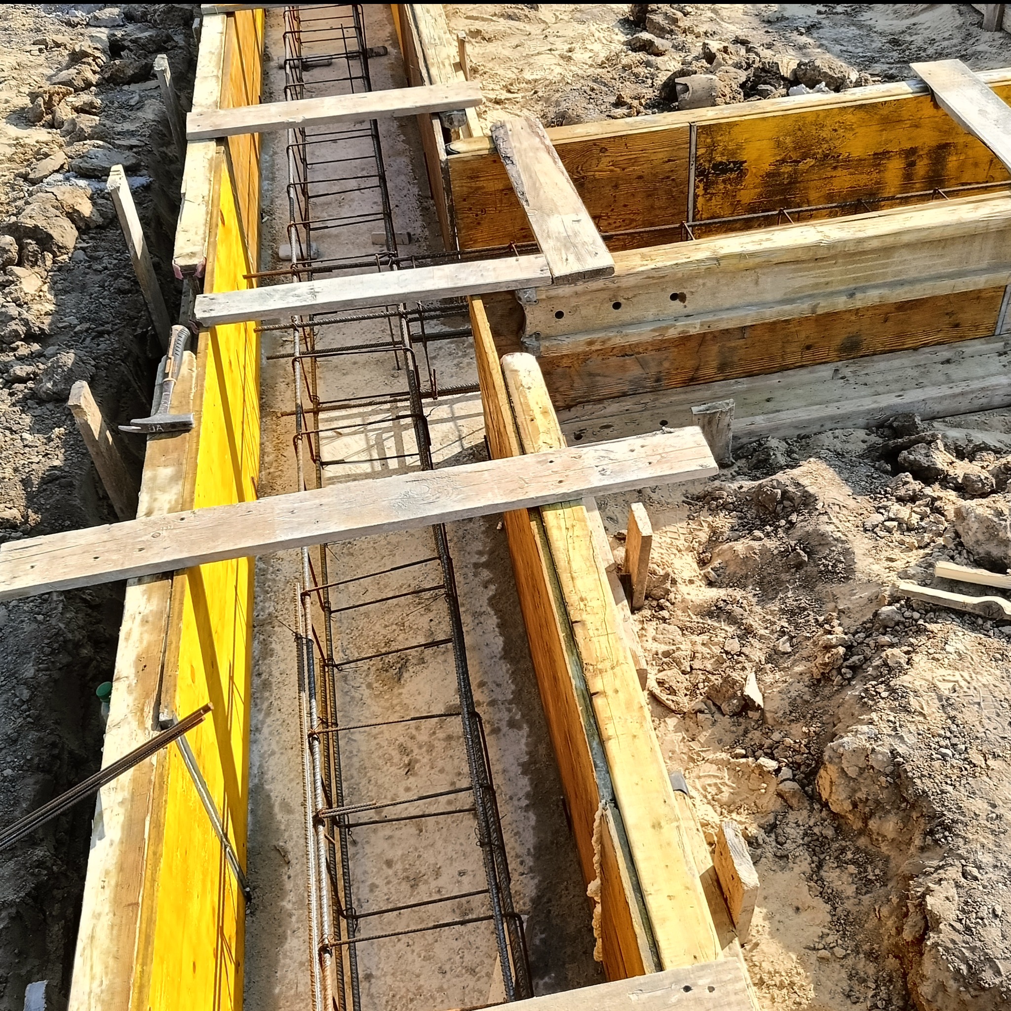 Szczegółowe ujęcie z góry na drewniany szalunek zbrojony stalą, przygotowany pod wylanie fundamentów. Żółte deski szalunkowe i ziemiste tło.