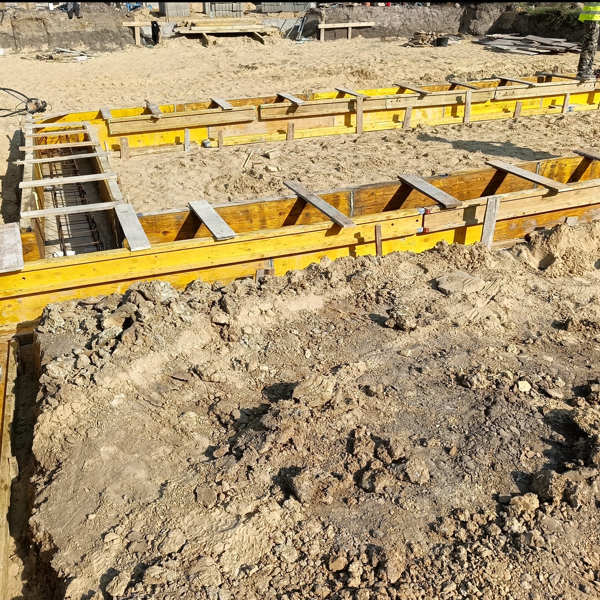 Żółte szalunki fundamentowe z widocznym zbrojeniem, przygotowane do zalania betonem. Widok z góry, na pierwszym planie ziemia.