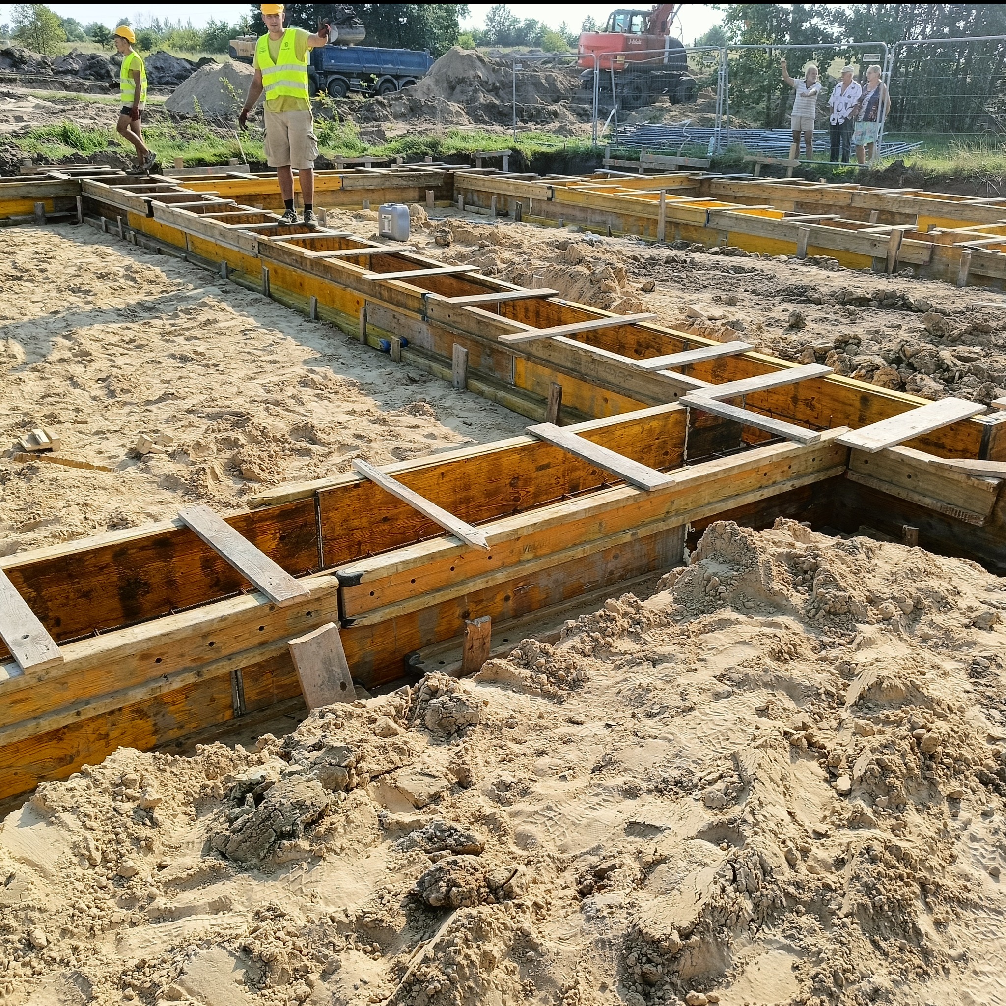 Wykopane fundamenty z drewnianym szalunkiem, widoczni pracownicy w odblaskowych kamizelkach i sprzęt budowlany w tle. Etap przygotowania do wylewania betonu.