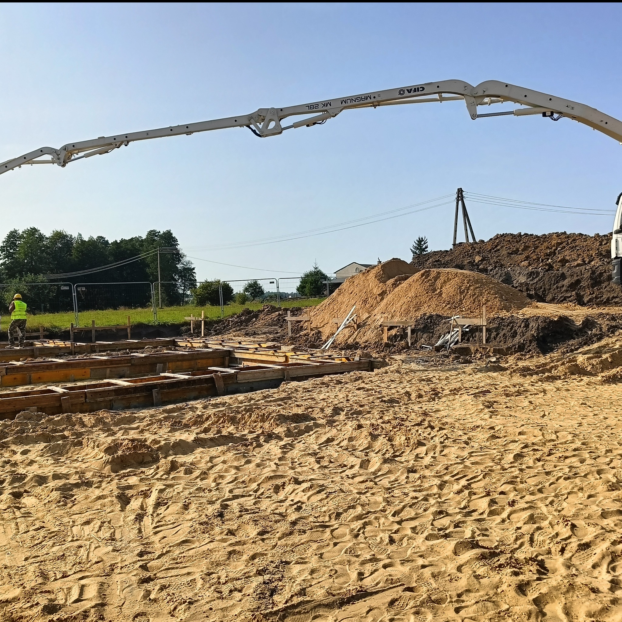 Wykopane fundamenty z szalunkiem i pracownikiem w kamizelce, widoczna pompa do betonu, hałda piachu i ziemi, teren budowy w słoneczny dzień.