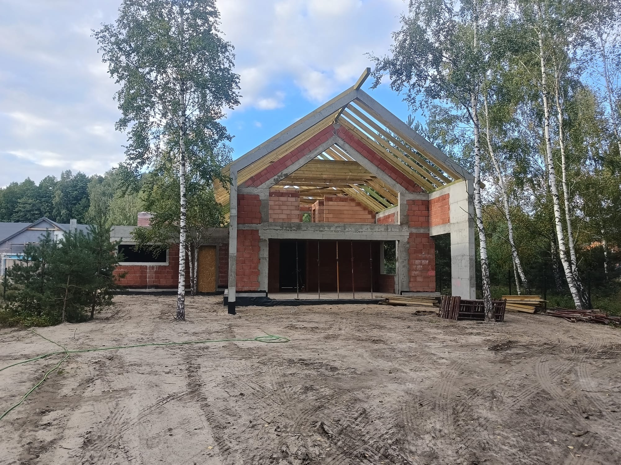 Surowy stan domu z czerwonej cegły z drewnianą konstrukcją dachu, otoczony drzewami brzozy i piaszczystym terenem w słoneczny dzień.