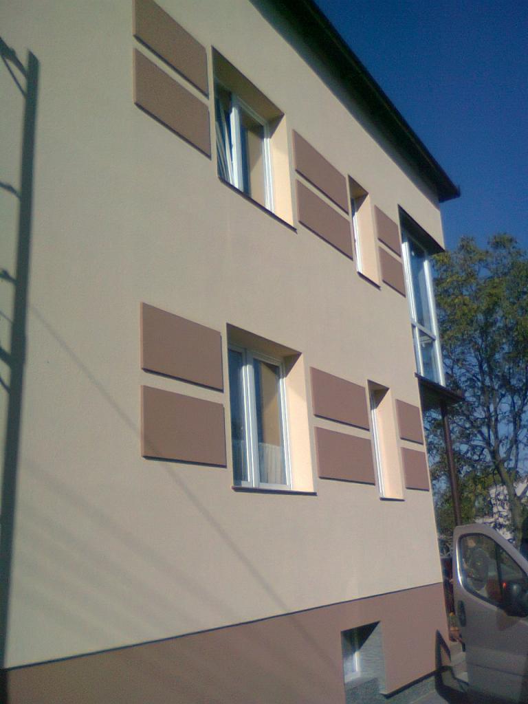 Ocieplony budynek z beżową elewacją i dekoracyjnymi, brązowymi panelami wokół okien, widoczny fragment samochodu dostawczego.