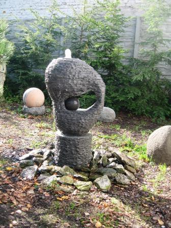 Nowoczesna fontanna ogrodowa z szarego łupka, składająca się z dwóch kul: mniejszej umieszczonej w otworze większej, z której wypływa woda, otoczona kamieniami i zielenią.