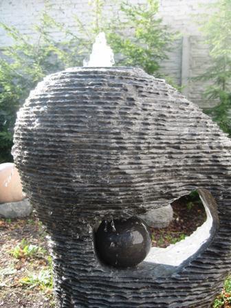 Nowoczesna fontanna ogrodowa z ciemnego kamienia o warstwowej strukturze, z wypływającą wodą i kulą w otworze, na tle zieleni i betonowego ogrodzenia.