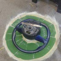 Niebieska kierownica samochodowa w trakcie naprawy, umieszczona na zielonej macie z włókna szklanego, widoczne narzędzie - szpachelka.