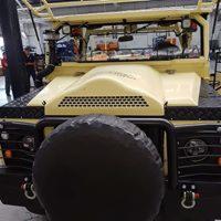 Żółty pojazd terenowy z czarnym kołem zapasowym z przodu, widoczny fragment dachu i wnętrza kabiny, z osobą w tle po lewej stronie.