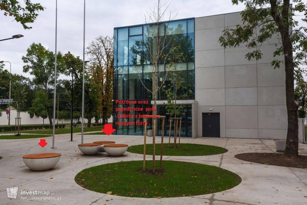 Nowoczesna przestrzeń publiczna z betonowymi siedziskami w kształcie mis, trawnikami w kształcie okręgów i budynkiem z dużą szklaną fasadą w tle.