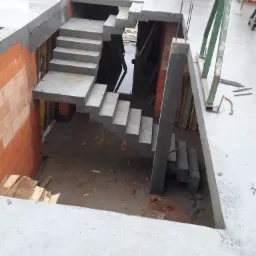 Surowe betonowe schody prowadzące w dół, w trakcie budowy, widoczne szalunki i cegły Porotherm na wilgotnym betonowym stropie.