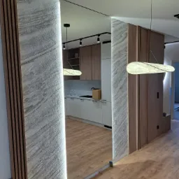 Nowoczesna aranżacja przedpokoju z lustrem wbudowanym w ścianę z dekoracyjną strukturą i drewnianymi lamelami. Widoczna kuchnia w tle i parkiet na podłodze.