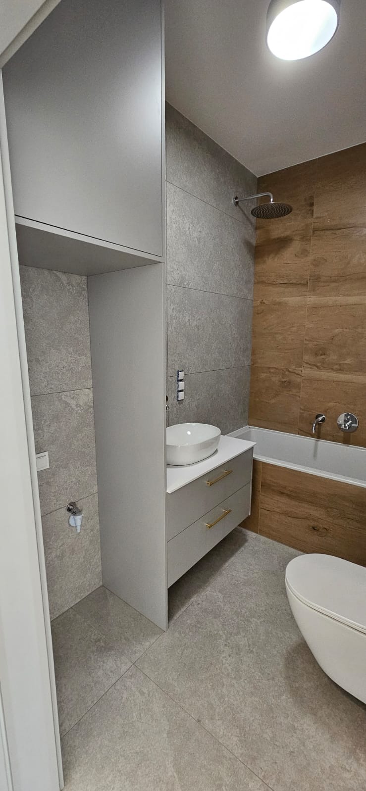 Nowoczesna łazienka z szarymi płytkami imitującymi kamień, drewnopodobnymi panelami przy wannie i białymi meblami z złotymi uchwytami, oświetlona okrągłą lampą sufitową.