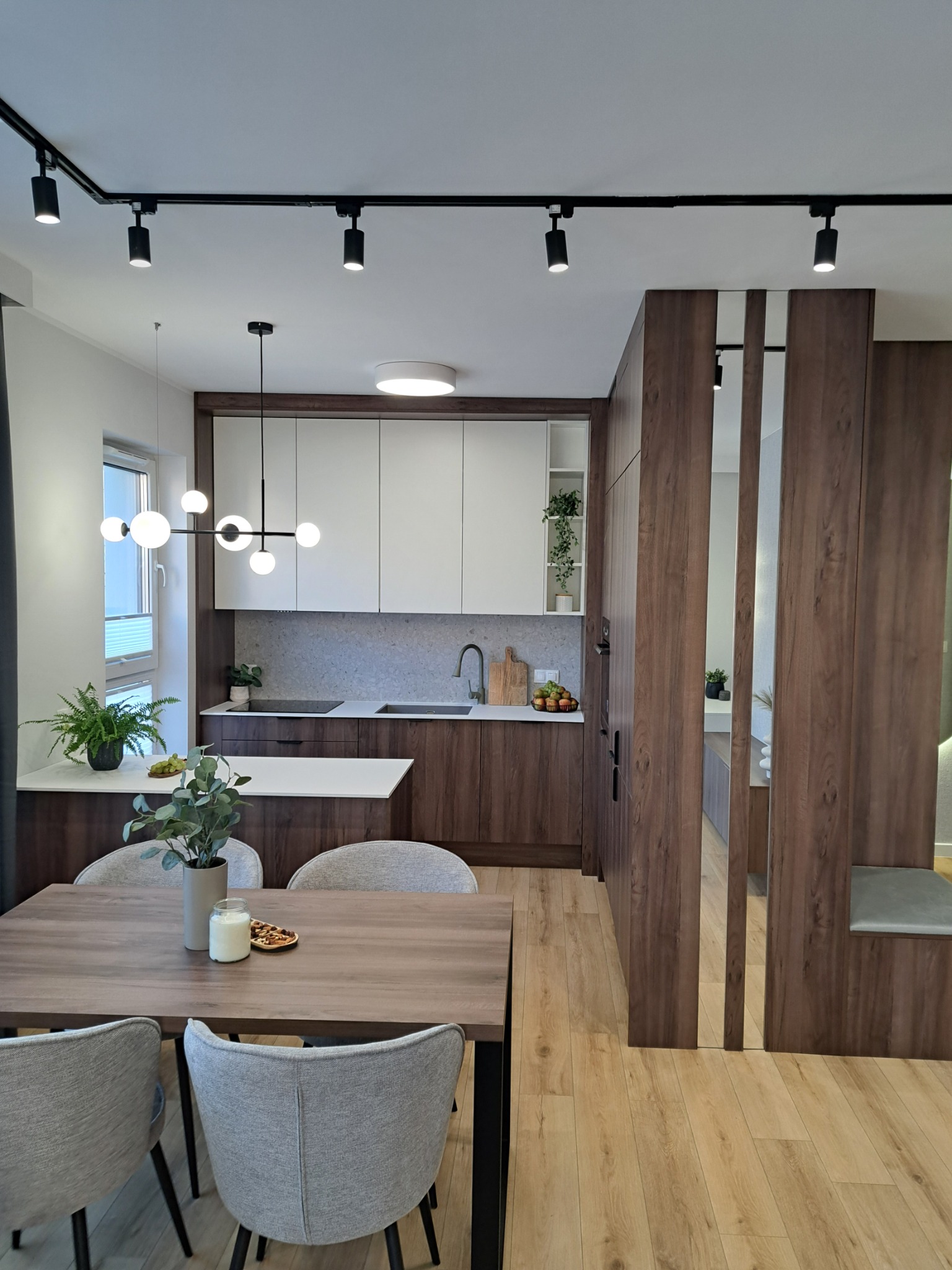 Nowoczesna aranżacja wnętrza: kuchnia z brązowo-białymi szafkami, drewnianym blatem i wyspą, oddzielona od jadalni drewnianą ścianką ażurową, oświetlona czarnymi reflektorami szynowymi i lampą...