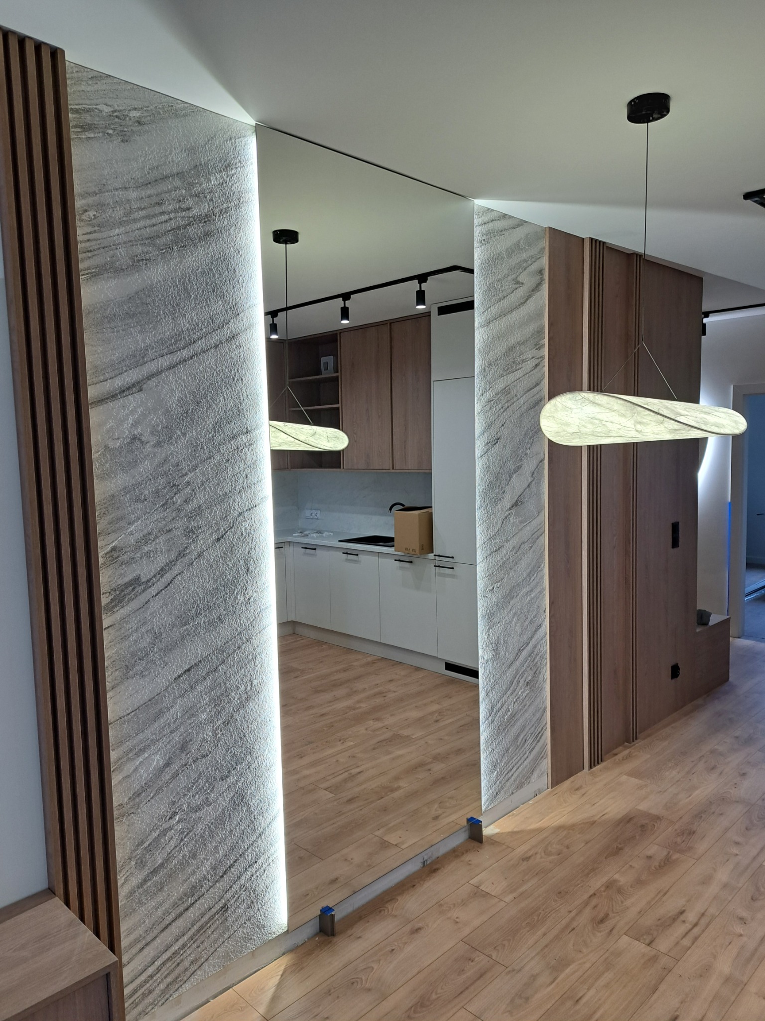 Nowoczesna aranżacja przedpokoju z lustrem wbudowanym w ścianę z dekoracyjną strukturą i drewnianymi lamelami. Widoczna kuchnia w tle i parkiet na podłodze.