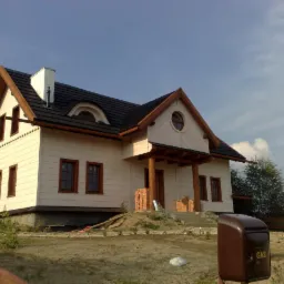 Jasny dom szkieletowy z ciemną dach&oacute;wką i drewnianymi elementami wykończeniowymi, widoczny z zewnątrz w słoneczny dzień. Na pierwszym planie skrzynka gazowa.