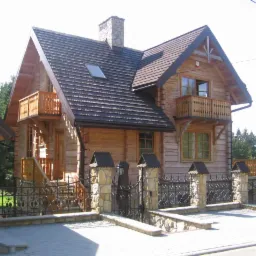 Dwukondygnacyjny dom z bali z balkonami, ciemnym dachem i kamiennym kominem. Ozdobne metalowe ogrodzenie z kamiennymi słupkami, kostka brukowa na podjeździe.