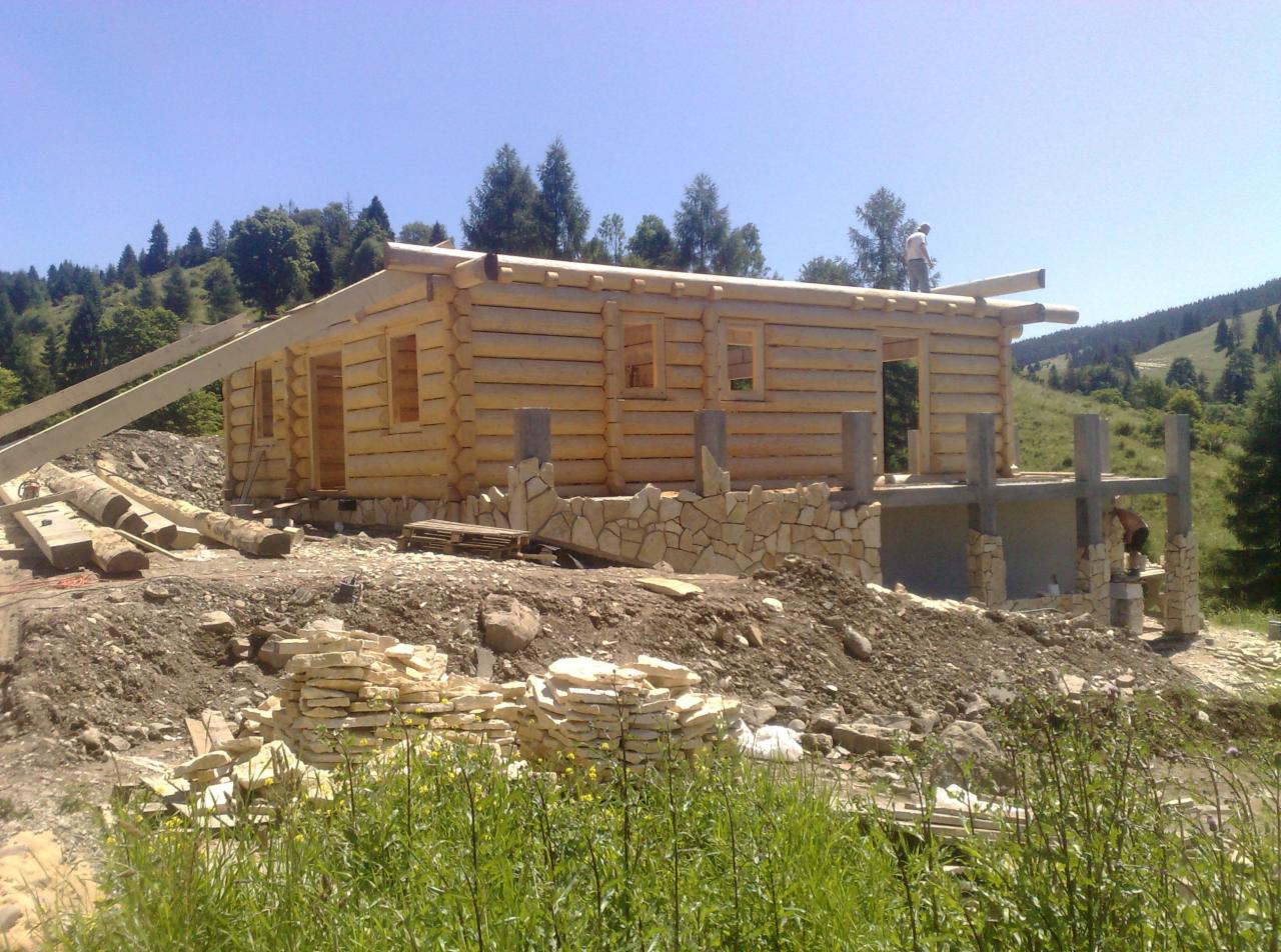 Wznoszenie domu z bali: surowa konstrukcja z drewnianych bali, kamienne fundamenty, widoczne elementy konstrukcyjne dachu i praca na wysokościach w otoczeniu zieleni.