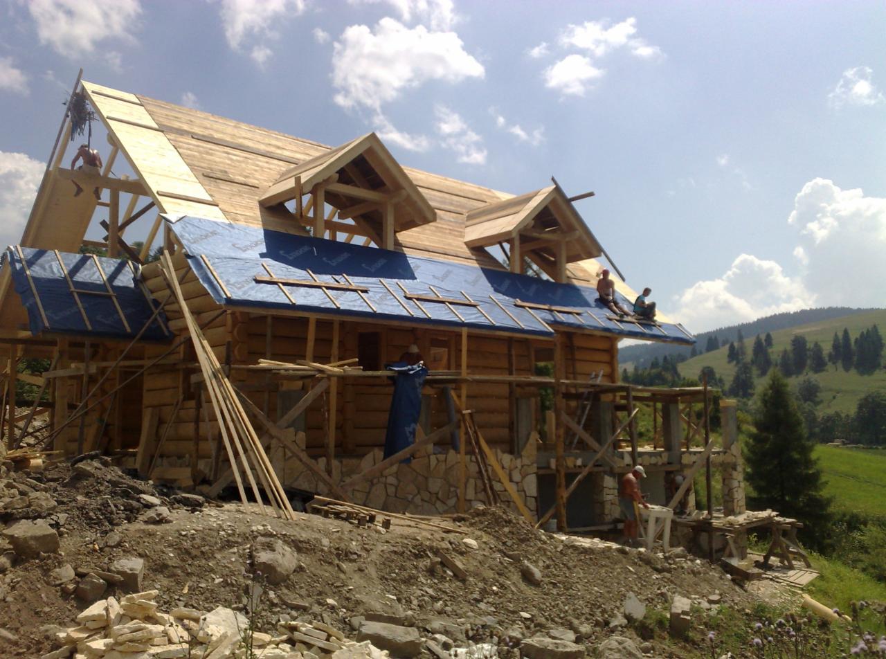 Budowa domu z bali: surowy stan z drewnianą konstrukcją dachu i ścian, częściowo obłożonych kamieniem. Na dachu pracownicy, w tle zielone wzgórza.