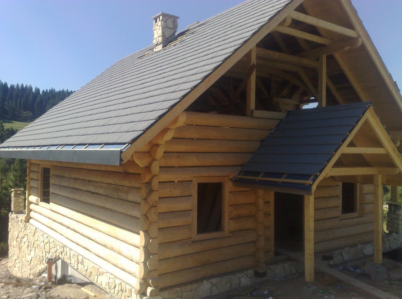 Surowy dom z bali z szarą dachówką, widoczny komin z kamienia i fundament. Konstrukcja w trakcie budowy, z elementami drewnianymi. W tle las iglasty.