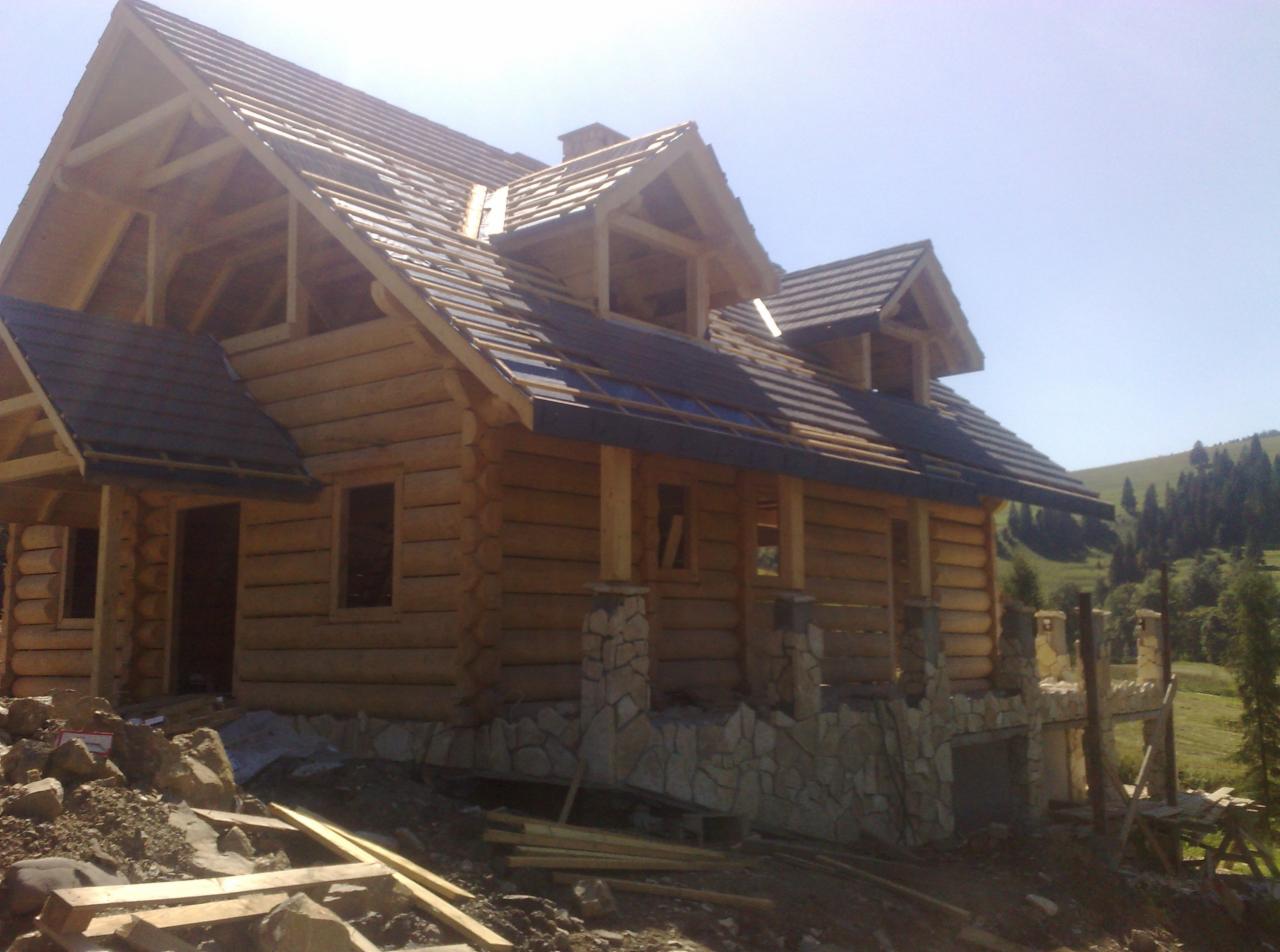 Surowy dom z bali z częściowo ułożonym dachem, kamienną podmurówką i widocznymi elementami konstrukcyjnymi na tle zielonego wzgórza.