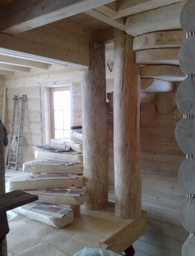 Wnętrze domu z bali w trakcie budowy: drewniane schody z zabezpieczeniami, okrągłe kolumny i ściany z bali, surowe drewno, wysokie pomieszczenie z oknem.