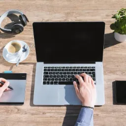 Osoba pracuje na laptopie z tabletem graficznym, kawa i słuchawki na biurku. Widok z góry, ciepłe światło.