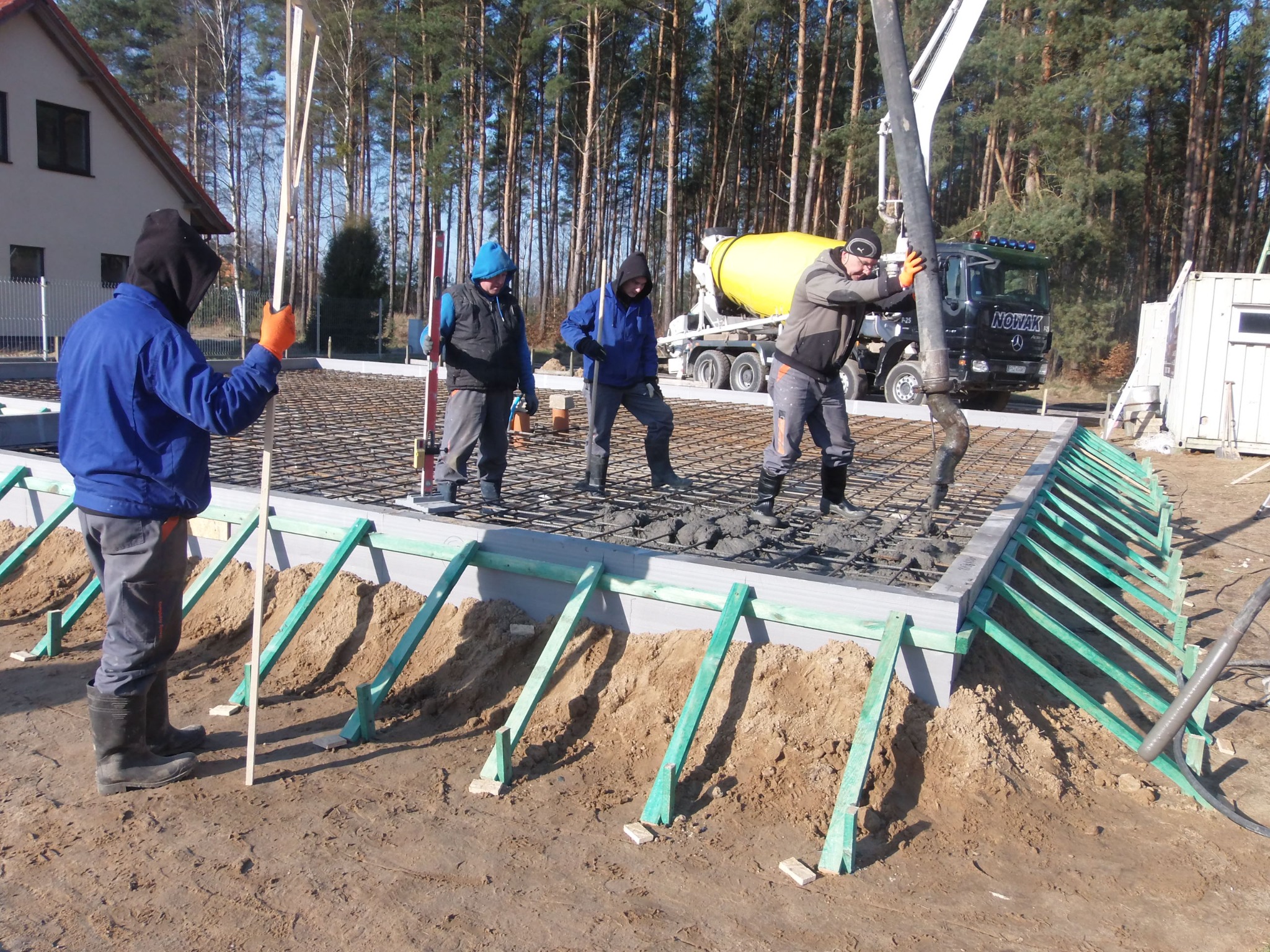 Wylewanie betonu na zbrojony fundament z użyciem pompy do betonu; widoczni robotnicy w roboczych ubraniach, szalunek z zielonych desek i częściowo wypełniona betonem siatka zbrojeniowa.