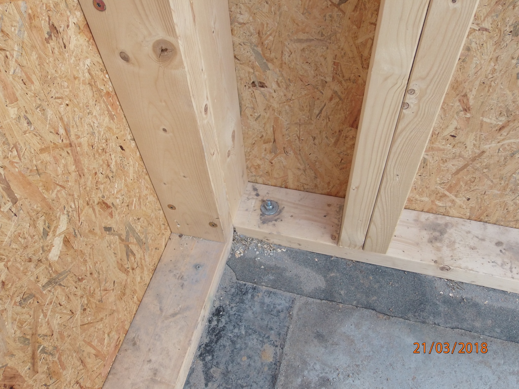 Fragment drewnianej konstrukcji szkieletowej budynku w trakcie budowy, widoczne płyty OSB i drewniane belki połączone śrubami, szara betonowa podłoga.