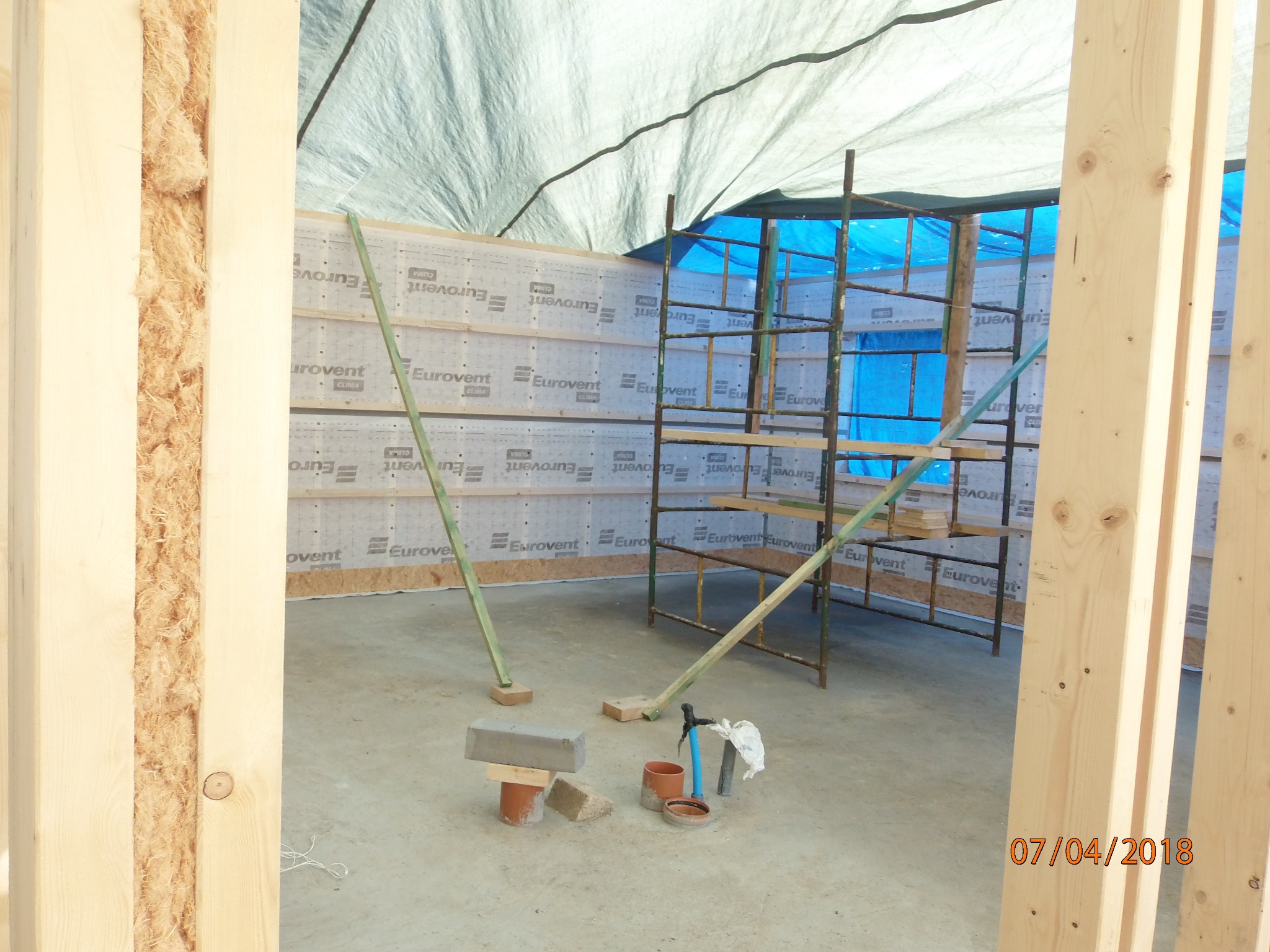 Wnętrze budynku w trakcie budowy: widoczne drewniane ramy ścian z izolacją, rusztowanie, folia ochronna, materiały budowlane i narzędzia na posadzce.