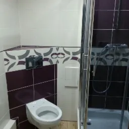 Nowoczesna łazienka z fioletowo-białymi płytkami, wiszącą toaletą i kabiną prysznicową ze srebrnymi profilami. Widoczne elementy dekoracyjne na płytkach.