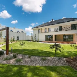 Nowoczesny, dwukondygnacyjny dom z płaskim dachem i balkonem, widoczny z perspektywy ogrodu z dekoracyjną pergolą i oddzielnym budynkiem gospodarczym w tle, Radomierzyce.