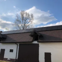 Brązowe panele słoneczne na dachu budynku gospodarczego z brązową dachówką, białe ściany, brązowe drzwi, błękitne niebo z chmurami i drzewo w tle.