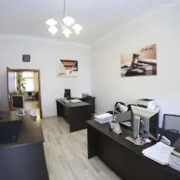 Jasne biuro z trzema biurkami, komputerami, lampą i plakatem z motywem prawnym na ścianie. Widoczny zegar ścienny i otwarte drzwi do kolejnego pomieszczenia.