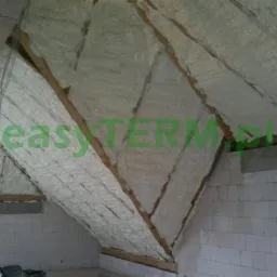 Poddasze z drewnianą konstrukcją krokwiową pokryte warstwą jasnej piany termoizolacyjnej, widoczne białe bloczki betonu komórkowego ścian i fragment okna.