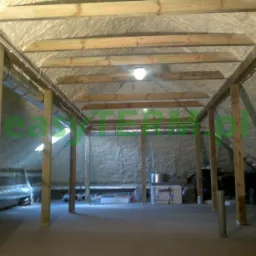 Poddasze po natrysku pianki izolacyjnej, widoczne drewniane belki konstrukcyjne i rury wentylacyjne, oświetlenie punktowe.