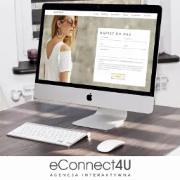 Ekran komputera iMac wyświetlający stronę internetową Ancoracollection.pl z formularzem kontaktowym, biała klawiatura i mysz na drewnianym biurku, w tle białe krzesło.