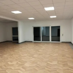 Jasne, przestronne wnętrze z parkietem ułożonym w jodełkę, białymi ścianami, podwieszanym sufitem z kwadratowymi lampami i dużymi przesuwnymi drzwiami balkonowymi.