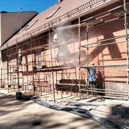 OS-BUD Ogólnobudowlane - Czyszczenie ceglanej elewacji budynku z rusztowania. Pracownicy w kaskach i roboczych ubraniach. Widoczne narzędzia i zabezpieczenia. Jasne, słoneczne światło.