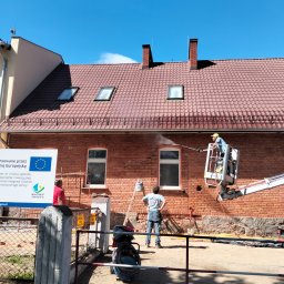 OS-BUD Ogólnobudowlane - Czyszczenie ceglanej elewacji budynku z podnośnika koszowego. Dwóch pracowników obserwuje proces. Widoczny fragment dachu z oknami i kominami.