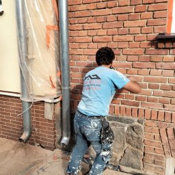 OS-BUD Ogólnobudowlane - Pracownik w trakcie spoinowania ceglanej elewacji budynku. Widoczne narzędzia i zabezpieczenia. Ujęcie z dołu, podkreślające detale muru i etap prac.