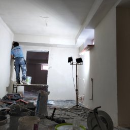 OS-BUD Ogólnobudowlane - Pracownik na podeście szpachluje ścianę w jasnym wnętrzu. Widoczne narzędzia, oświetlenie budowlane i surowe wykończenie. Ujęcie z dołu, podkreślające etap prac.