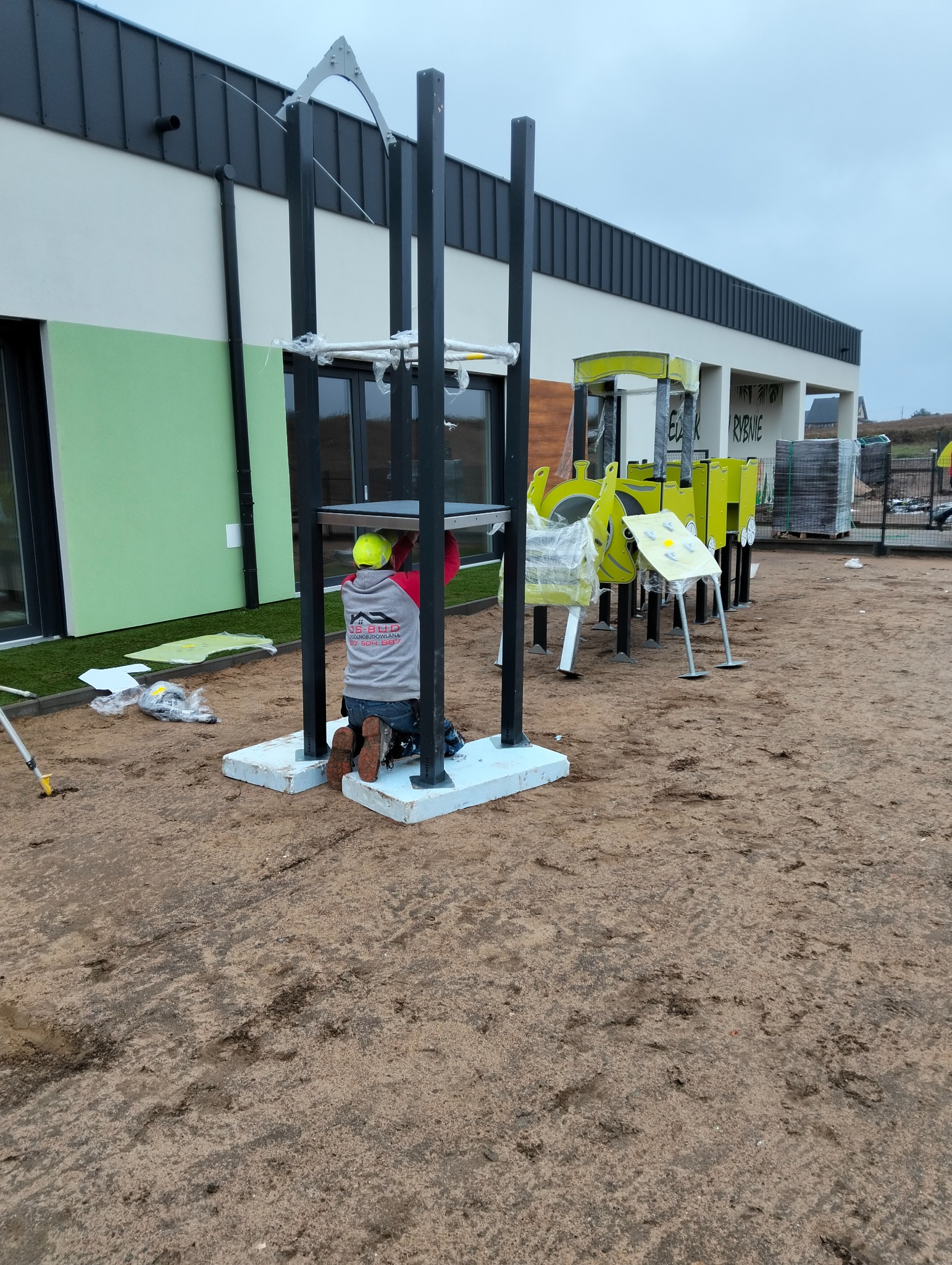 Pracownik w kasku montuje element placu zabaw na betonowej podstawie; w tle budynek z elewacją w odcieniach bieli, zieleni i szarości.