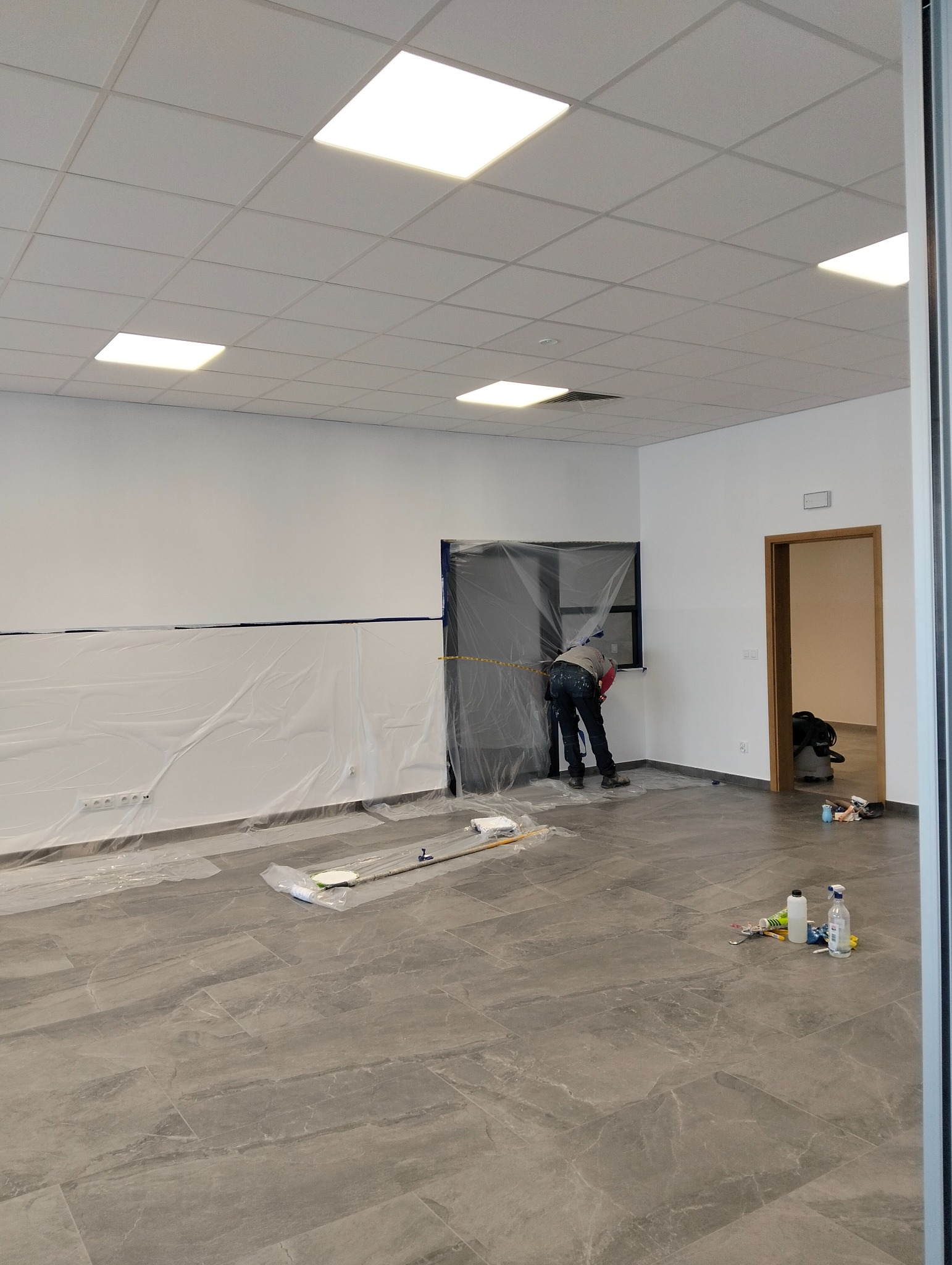 Przygotowanie do malowania wnętrza: zabezpieczone folią ściany i okno, widoczny pracownik przy oknie, na podłodze narzędzia i akcesoria malarskie, szare płytki podłogowe, widok na otwarte drzwi...