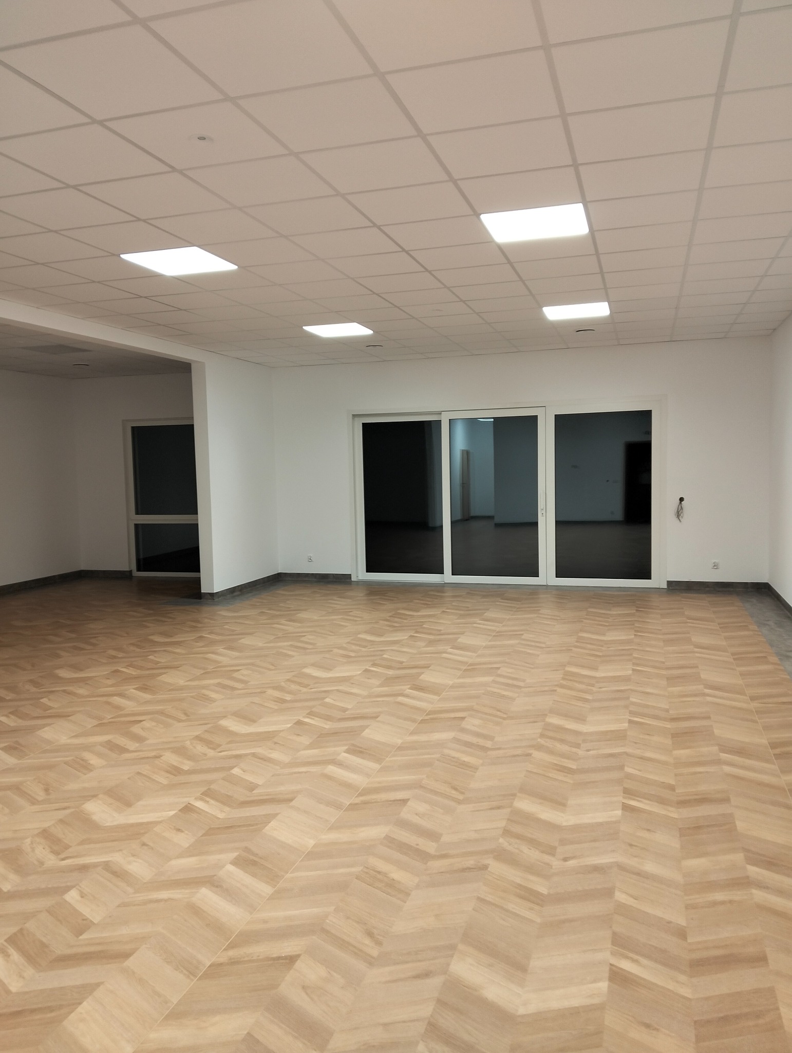 Jasne, przestronne wnętrze z parkietem ułożonym w jodełkę, białymi ścianami, podwieszanym sufitem z kwadratowymi lampami i dużymi przesuwnymi drzwiami balkonowymi.