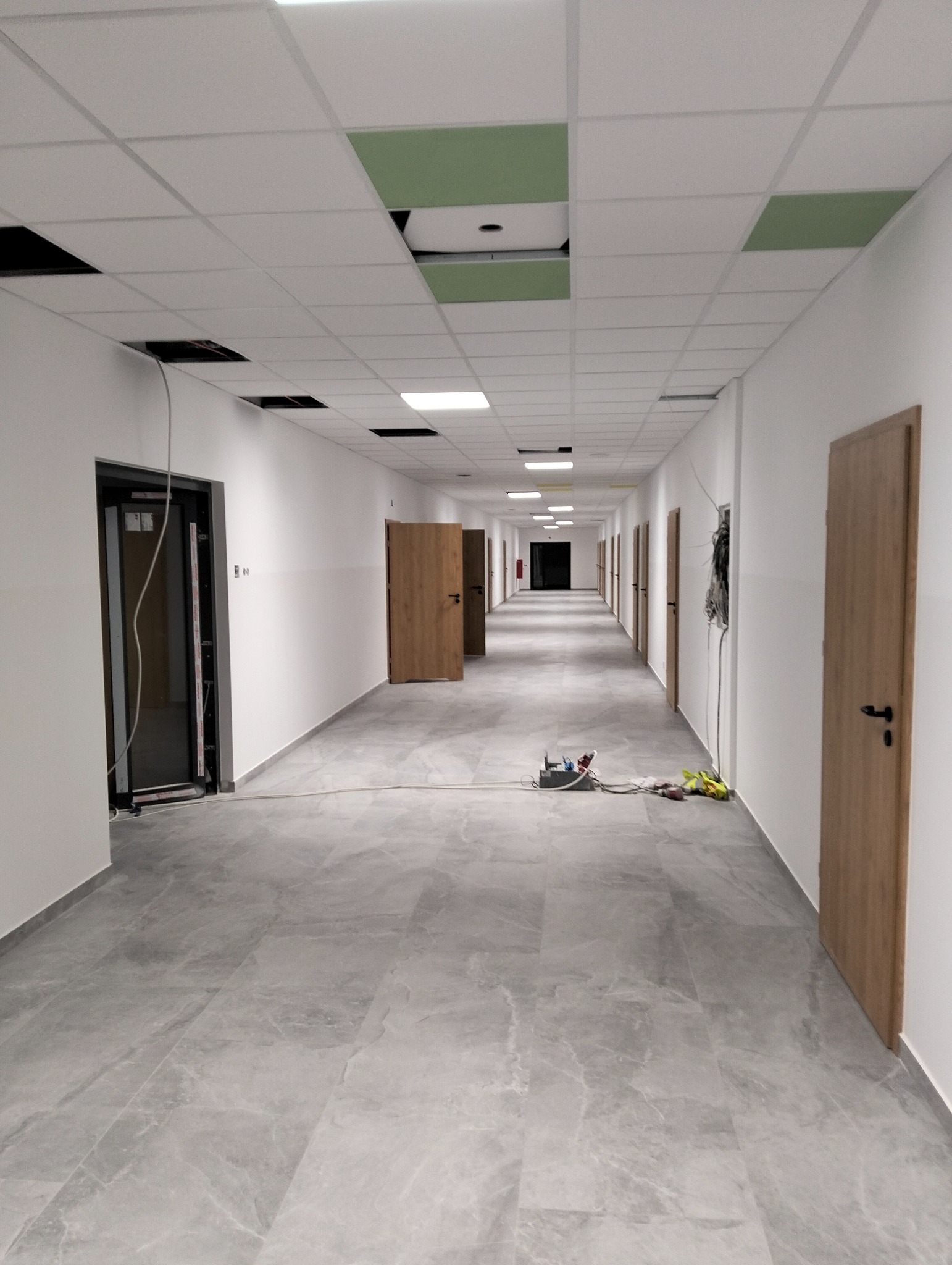 Długi korytarz w Rybnie z szarą marmurkową podłogą, białymi ścianami, drewnianymi drzwiami i podwieszanym sufitem z otworami na oświetlenie oraz narzędzia na podłodze wskazujące na trwające prace...