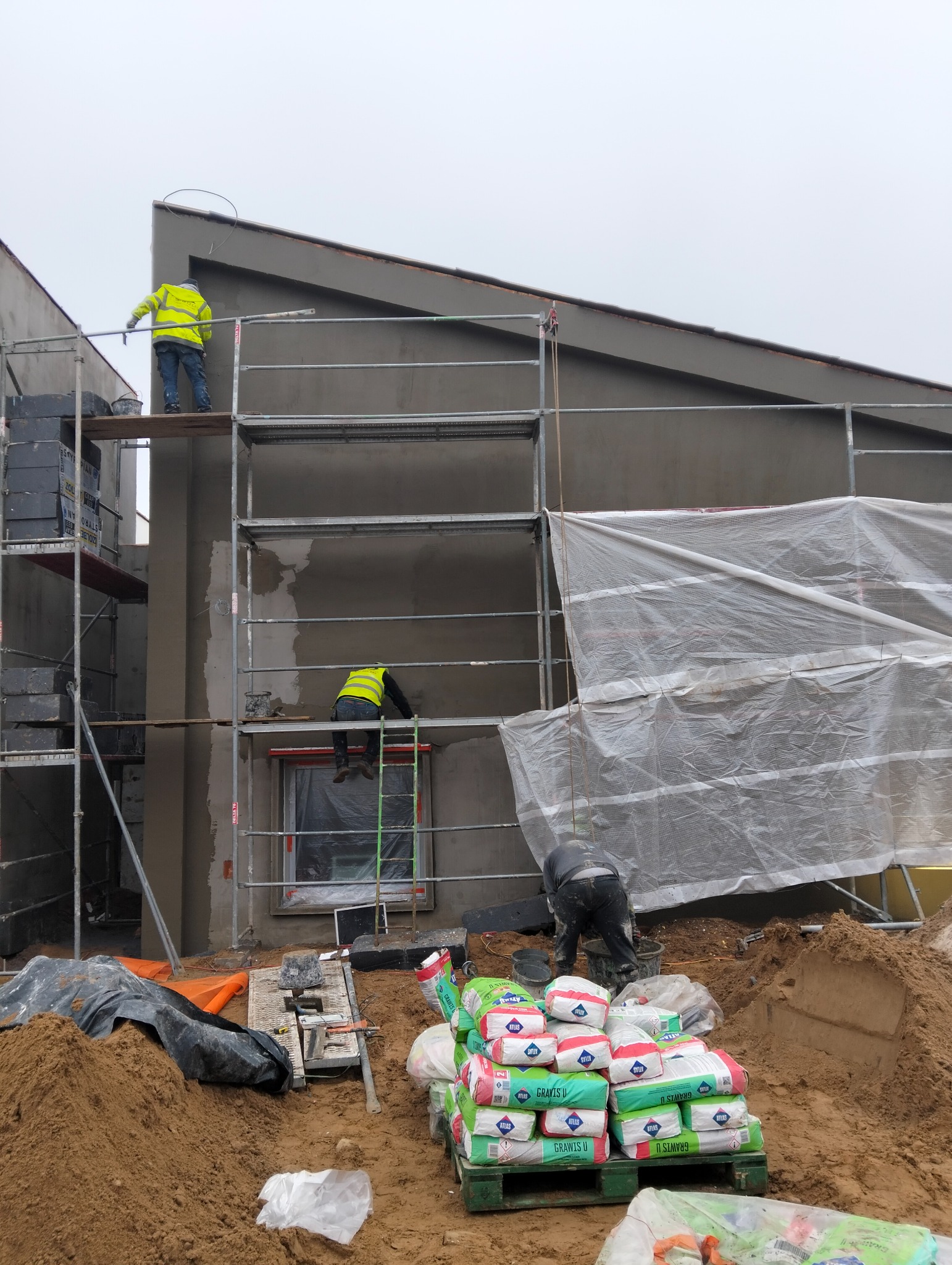 Pracownicy na rusztowaniu podczas nakładania tynku na elewację domu. Widoczne worki z zaprawą Atlas GraviS U i narzędzia budowlane.