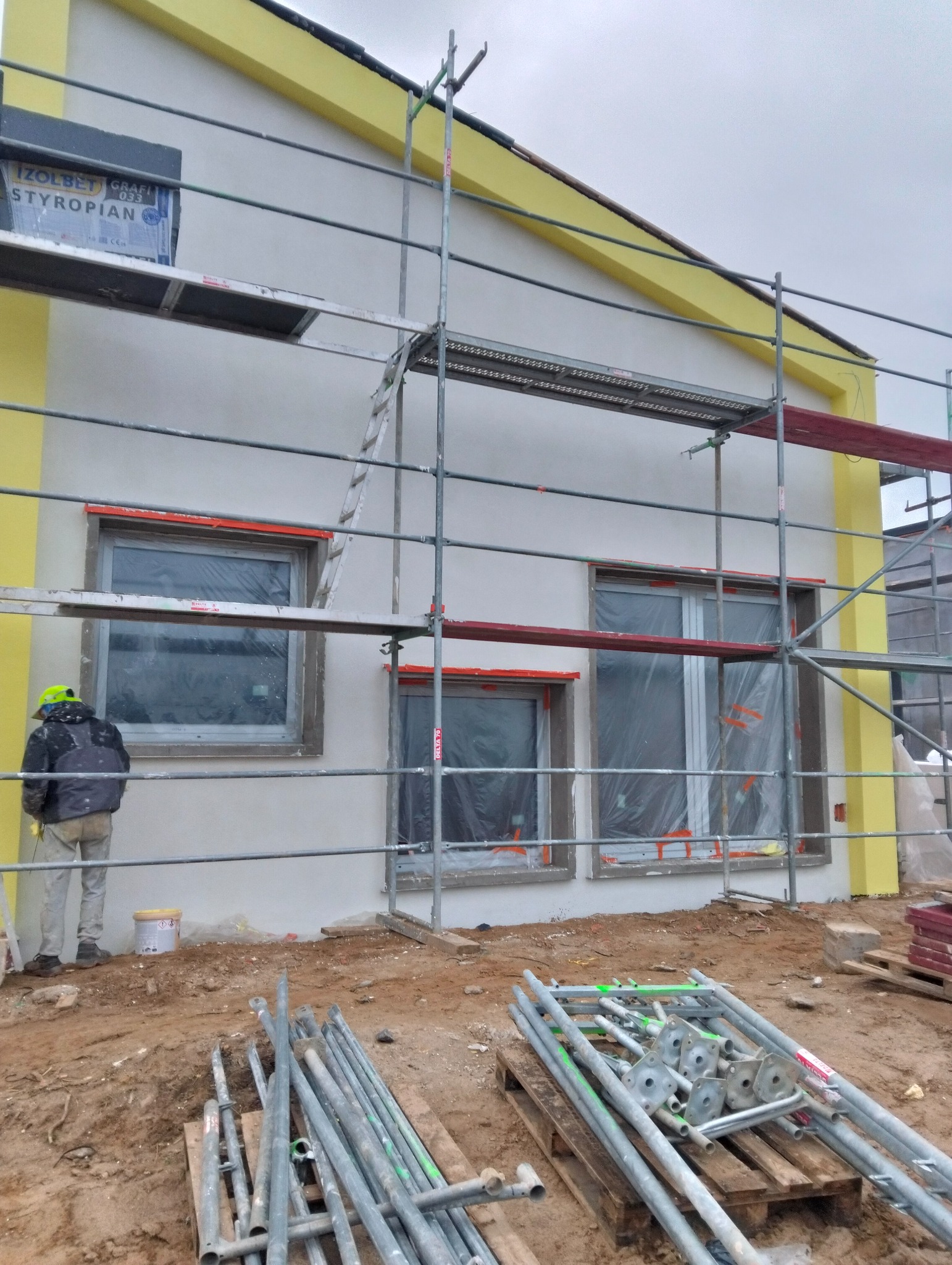 Ocieplony dom w trakcie prac elewacyjnych. Widoczne rusztowania, okna zabezpieczone folią, pracownik w kasku i materiały budowlane na ziemi. Żółte akcenty kolorystyczne.