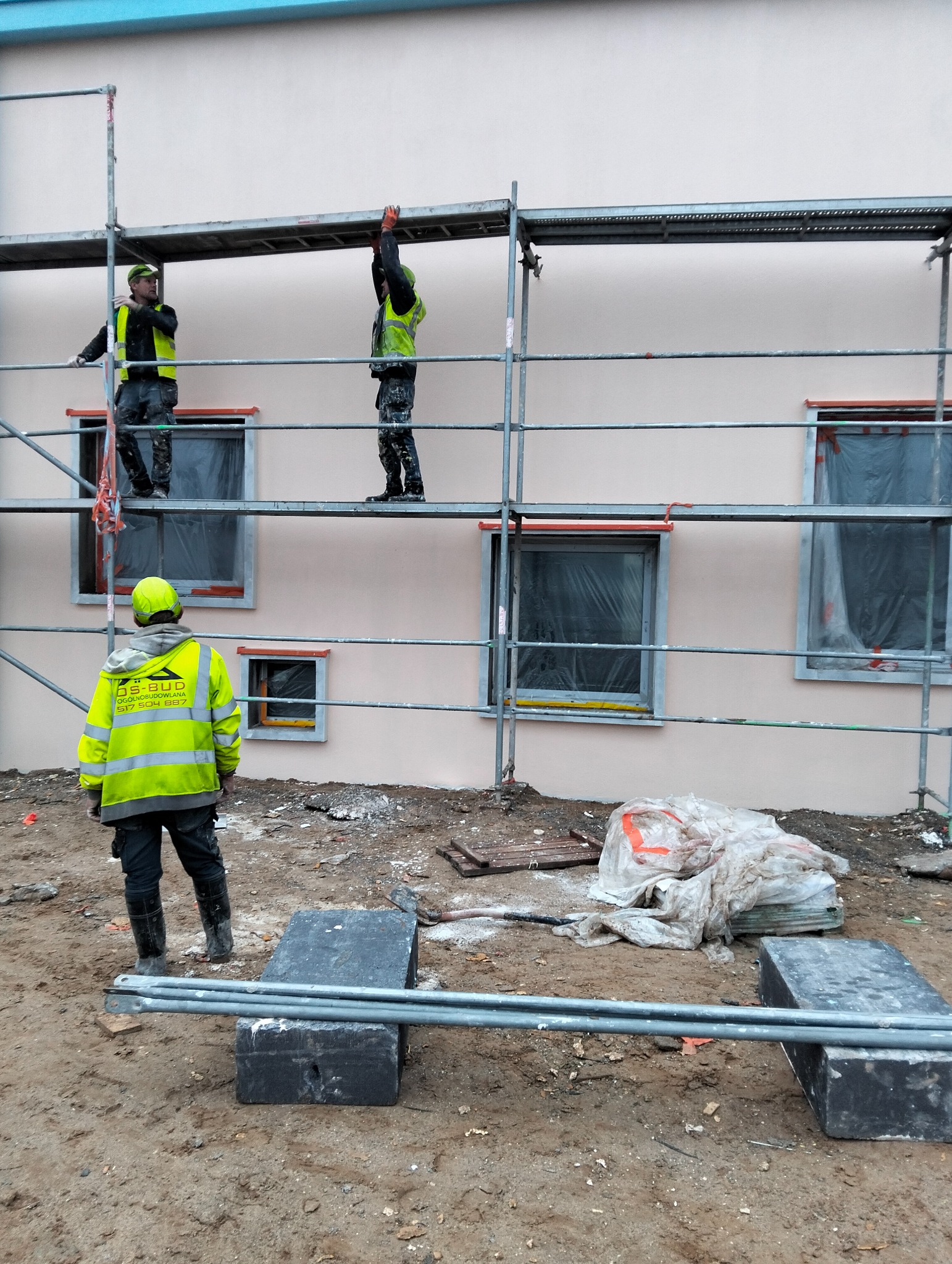 Pracownicy na rusztowaniu podczas prac elewacyjnych. Widoczne okna zabezpieczone folią i fragment otynkowanej ściany w odcieniu beżu. Jeden z pracowników patrzy w dół.