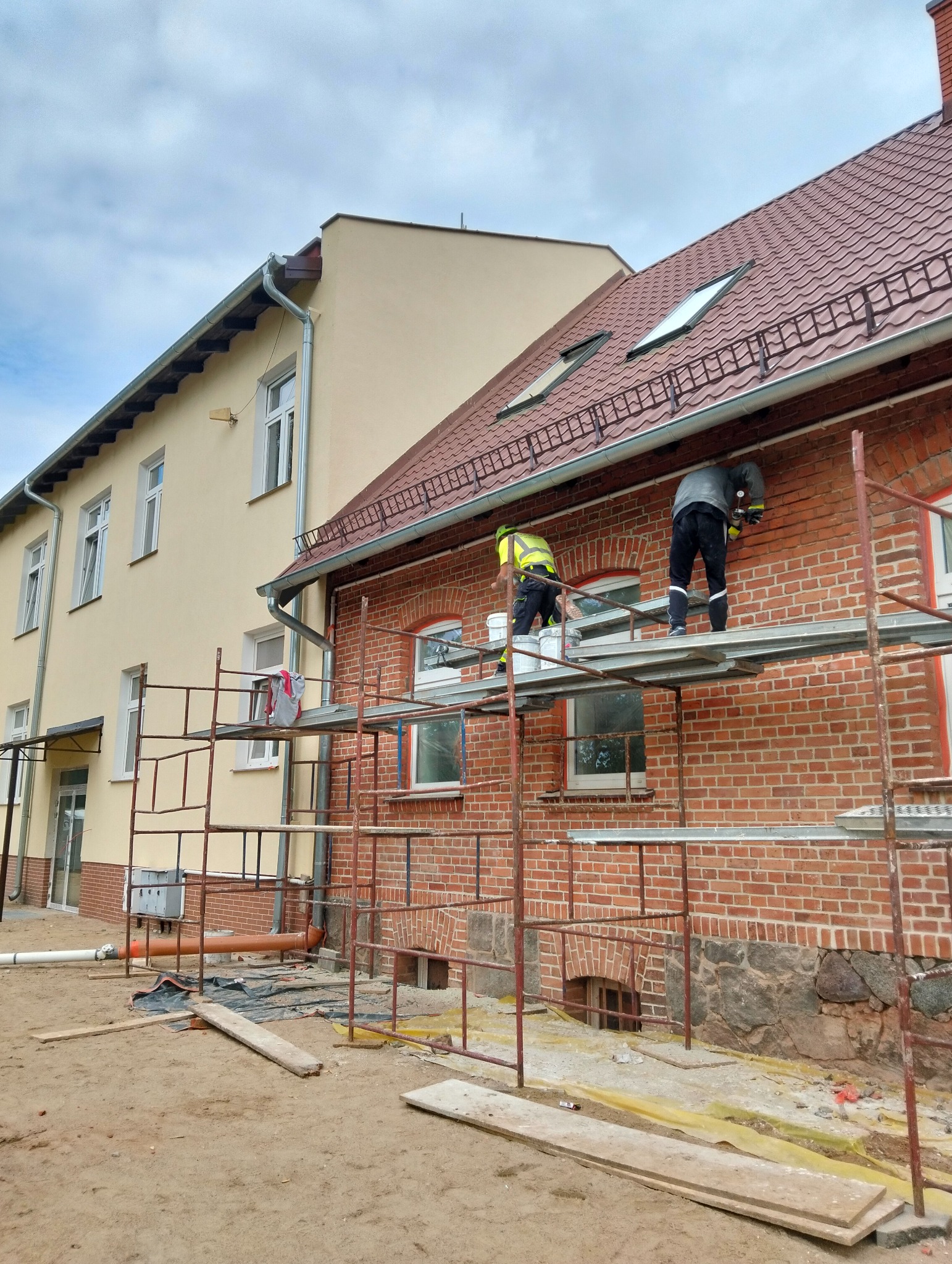 Dwóch pracowników na rusztowaniu przy ceglanym budynku, prawdopodobnie podczas prac elewacyjnych. Widoczne narzędzia i materiały budowlane. Budynek z czerwonej cegły.