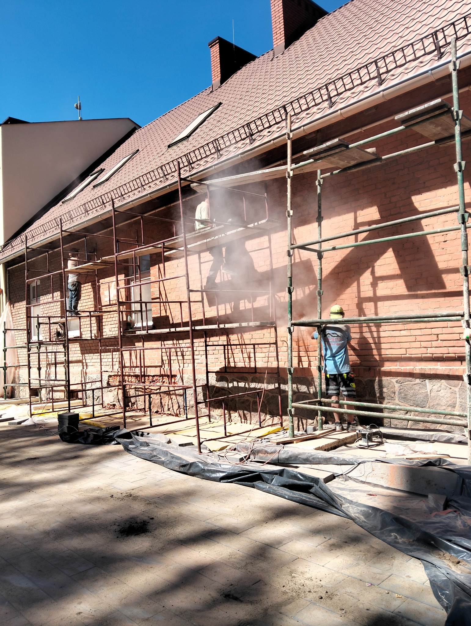 Czyszczenie ceglanej elewacji budynku z rusztowania. Pracownicy w kaskach i roboczych ubraniach. Widoczne narzędzia i zabezpieczenia. Jasne, słoneczne światło.