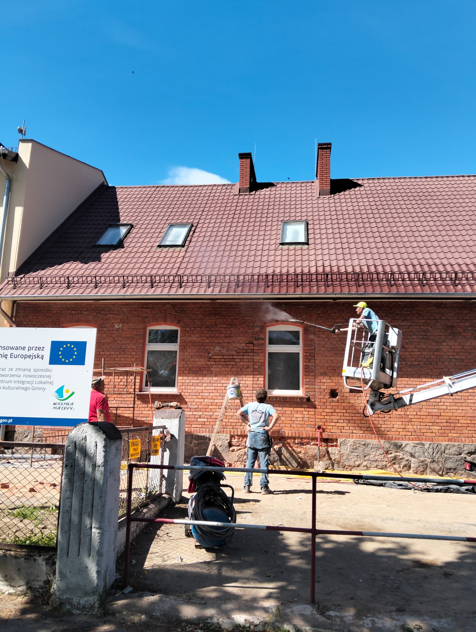 Czyszczenie ceglanej elewacji budynku z podnośnika koszowego. Dwóch pracowników obserwuje proces. Widoczny fragment dachu z oknami i kominami.